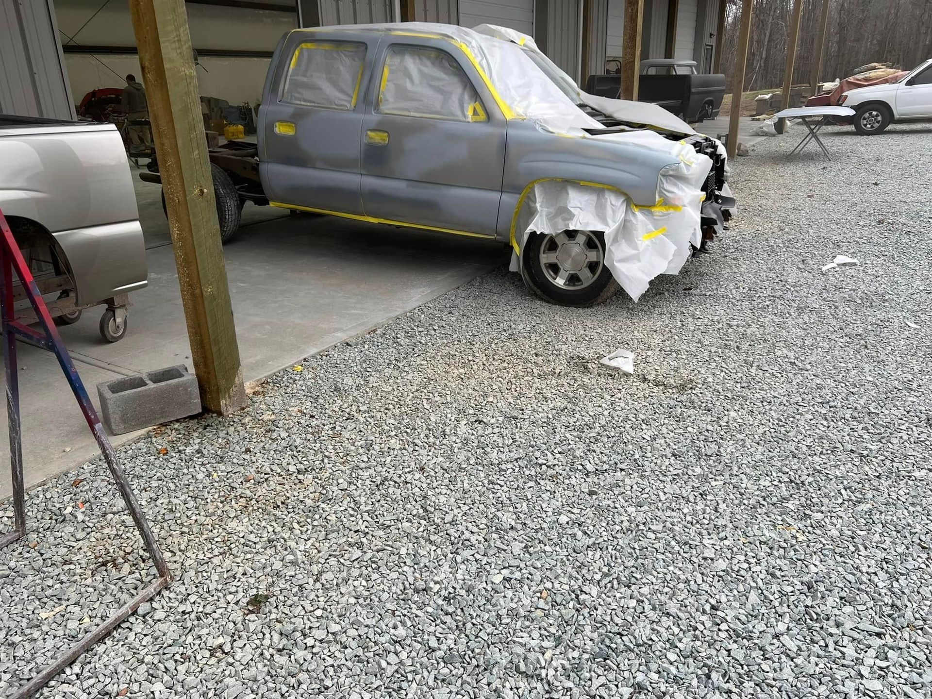 Gray car covered in primer and masking tape, parked in a gravel yard near a shed