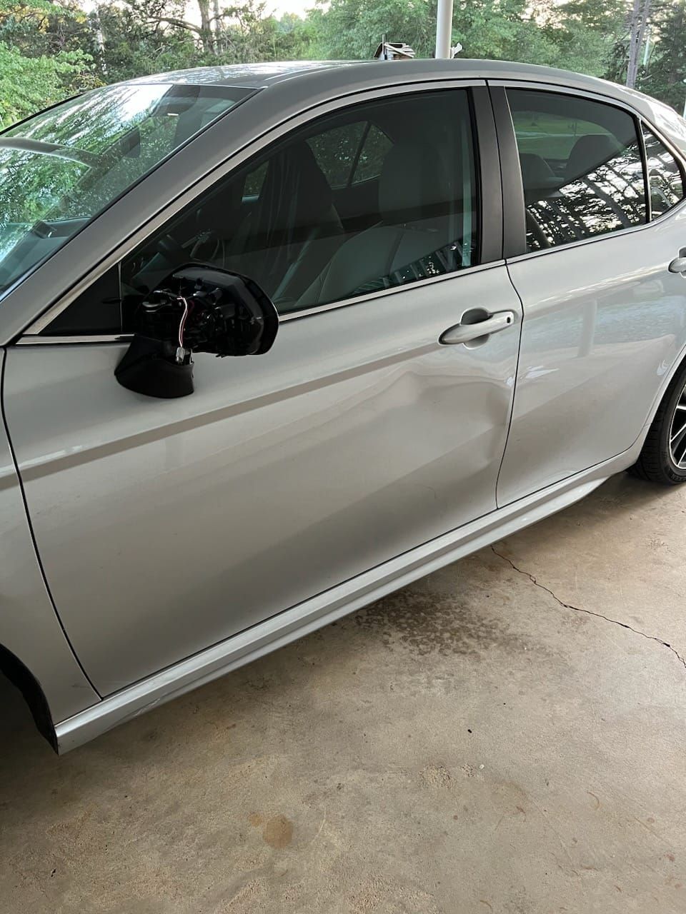 Silver sedan parked in a driveway, showing visible dents on the side door and side mirror.
