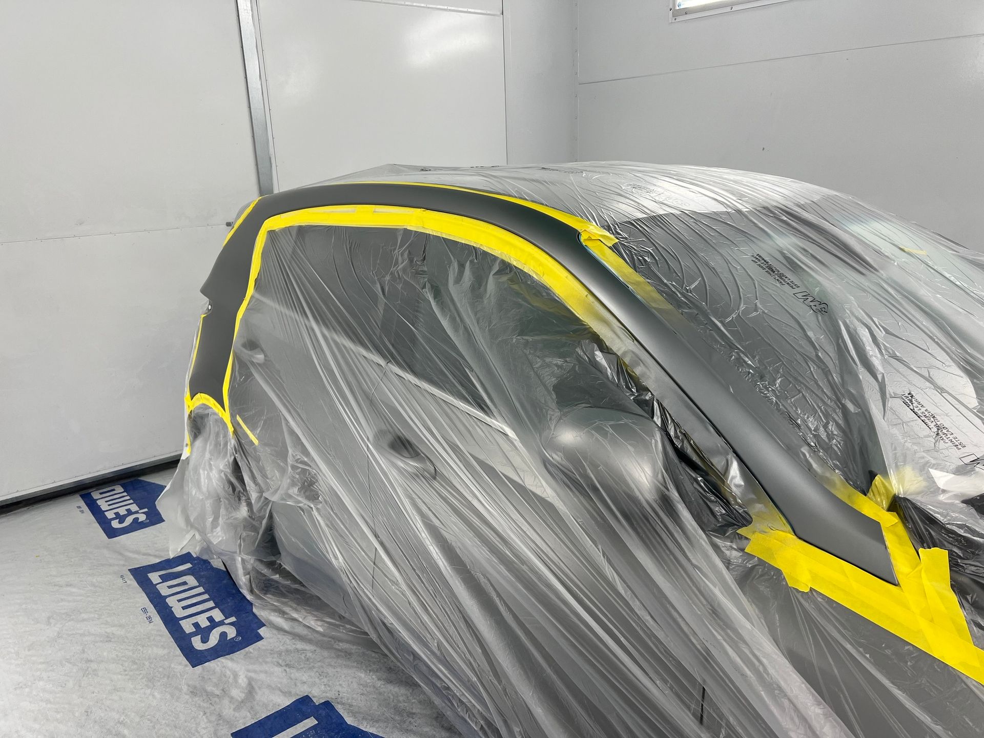 Car masked in clear plastic and yellow tape inside a paint booth, likely being prepped for painting