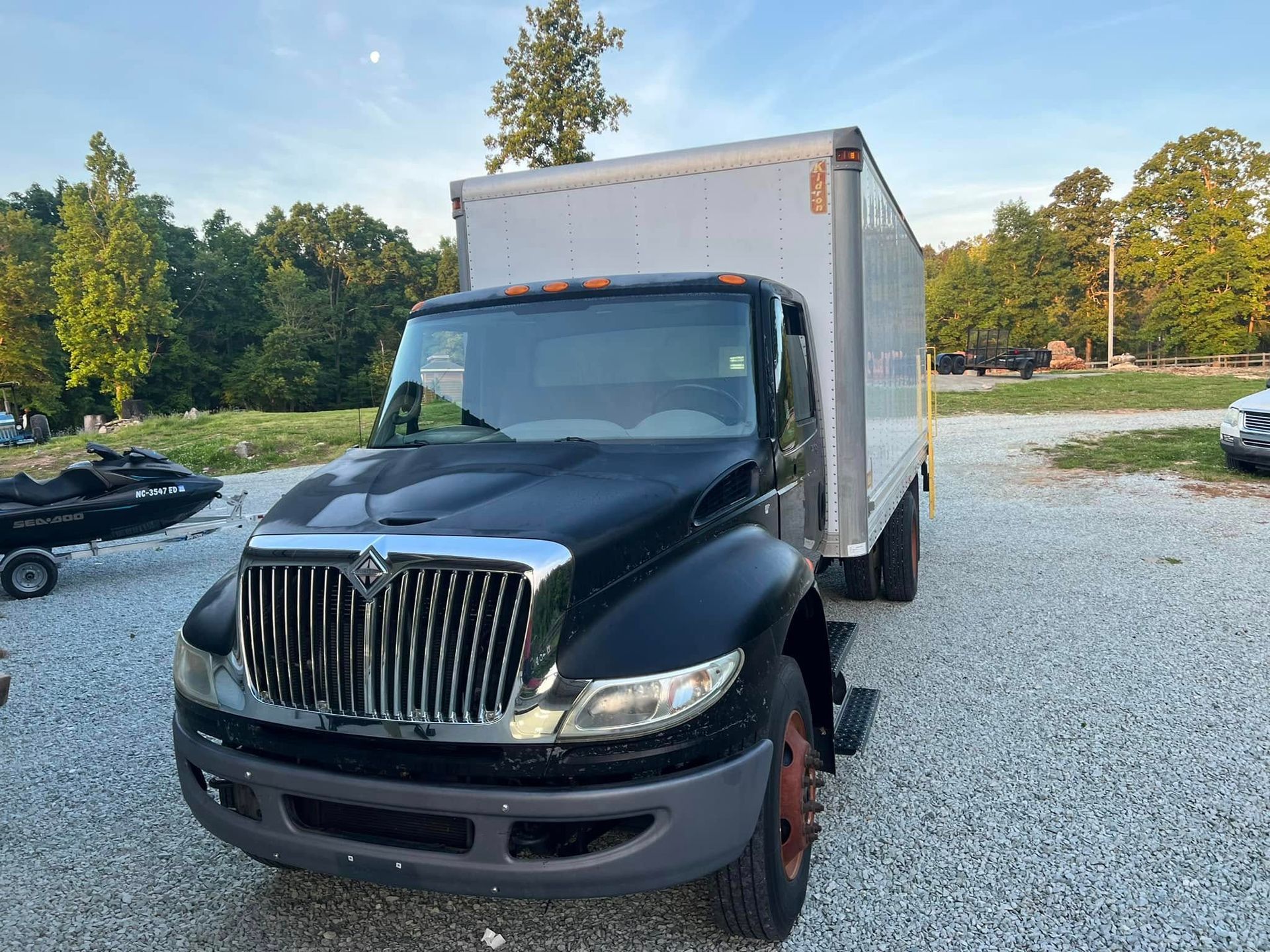 Black box truck parked outdoors on a gravel lot, with trees and a car in the background.