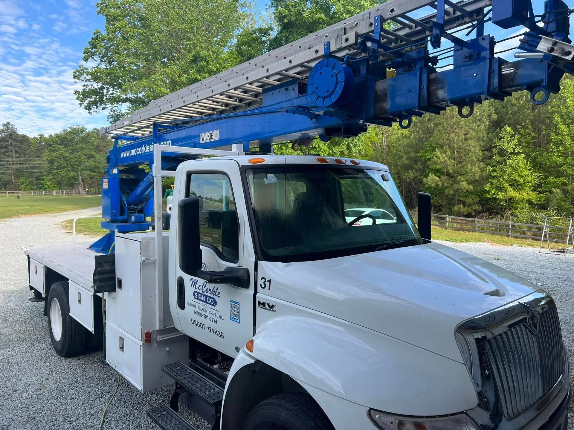 White utility truck with blue boom lift parked on a roadside, trees and blue sky in the background