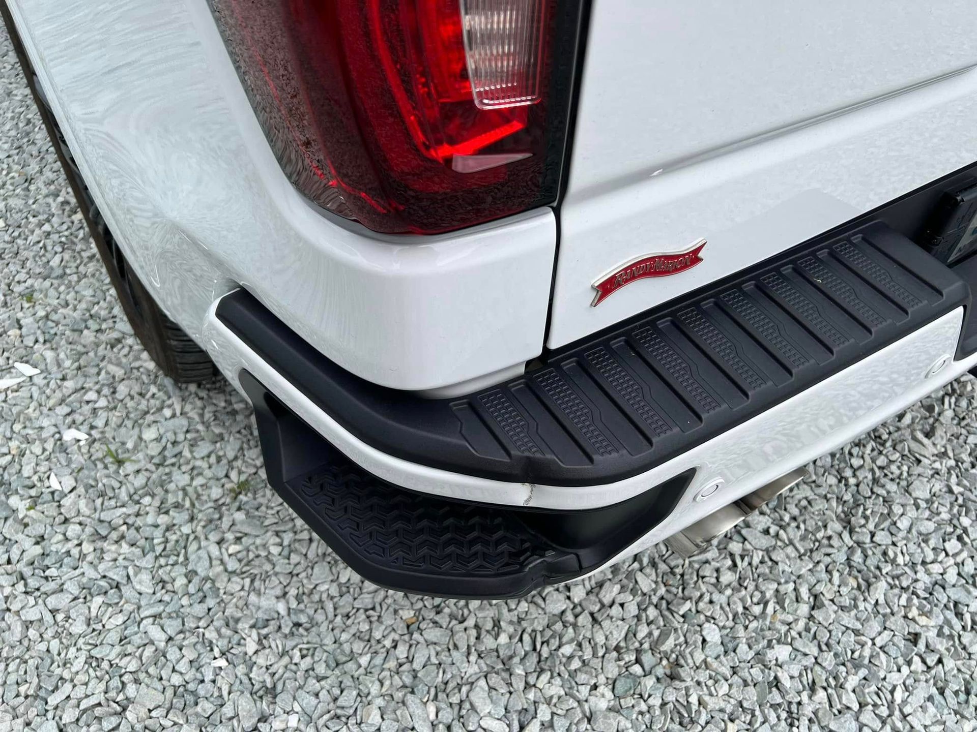 Rear corner of a white pickup truck with red taillight and black step bumper near gravel driveway