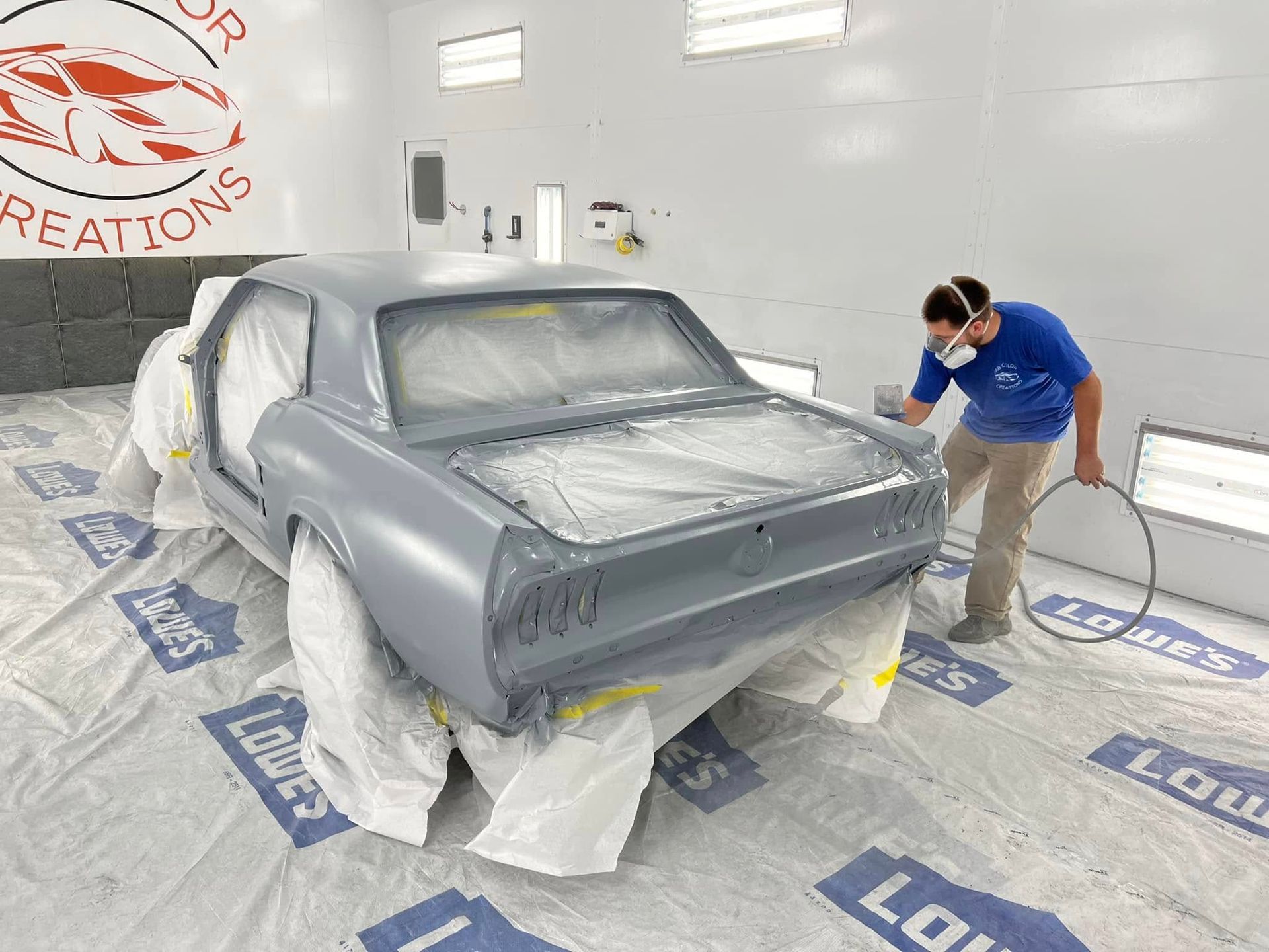 Person spray-paints a gray classic car body in a bright auto body shop.