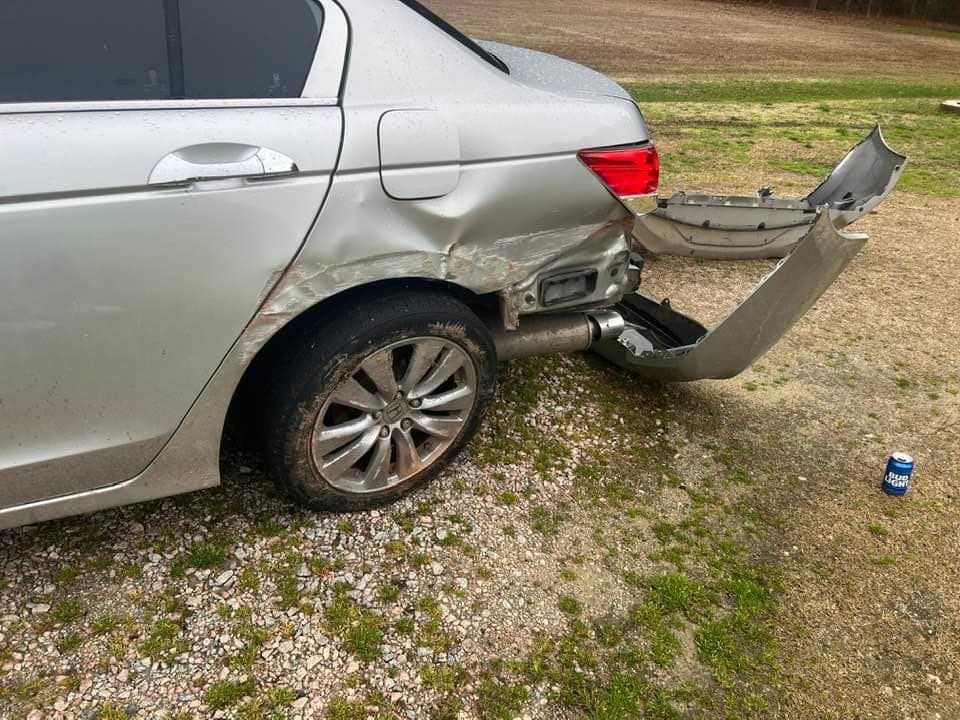Damaged silver sedan with rear-end collision and detached bumper on gravel roadside