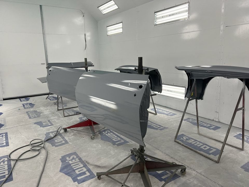 Car parts in a white spray booth, including a silver panel and two black components on stands.