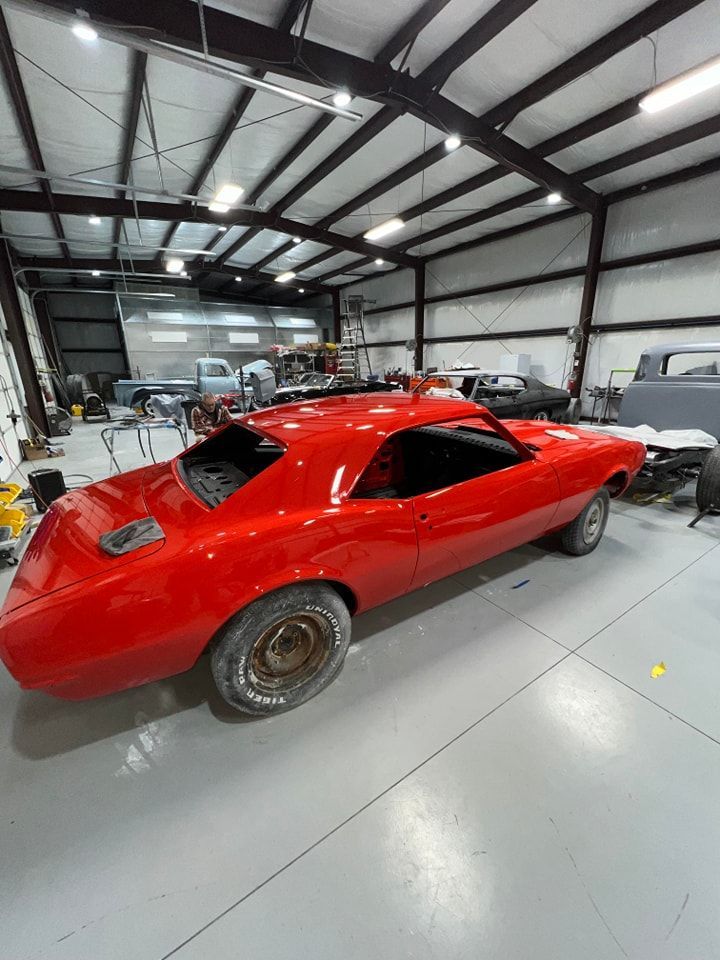 Red classic muscle car in a garage workshop under bright overhead lights.