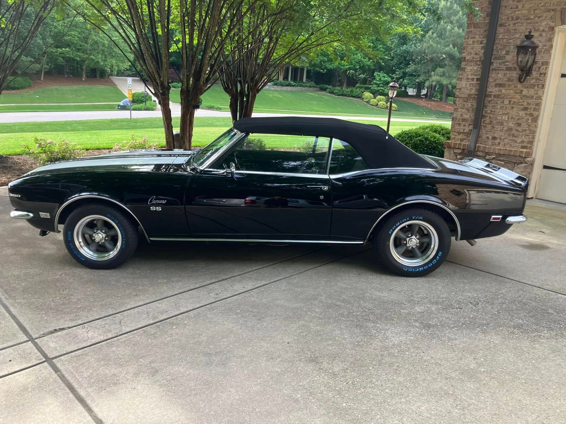 Black classic car parked on a driveway beside a brick house with green trees in the background
