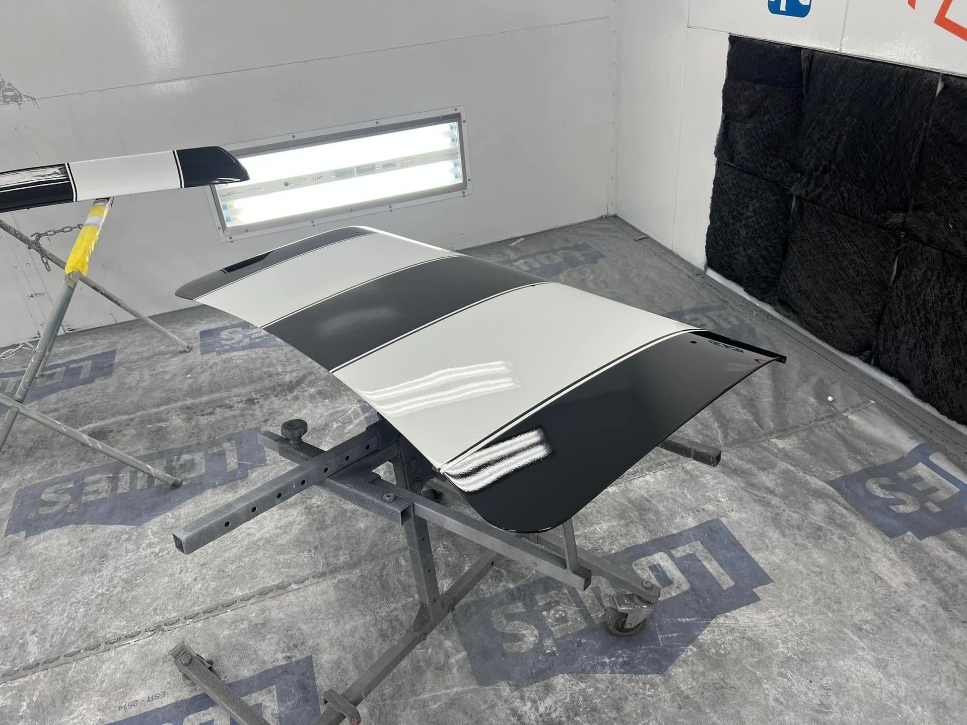 Black-and-white car hood on a stand in a workshop with bright lights and protective floor covering