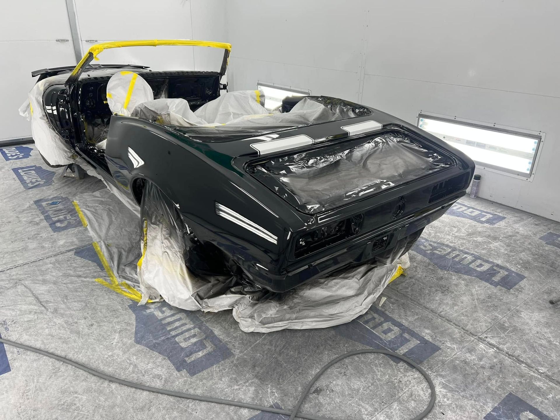 Black convertible body in an auto body shop, wrapped in plastic and tape, with yellow masking on the windshield.