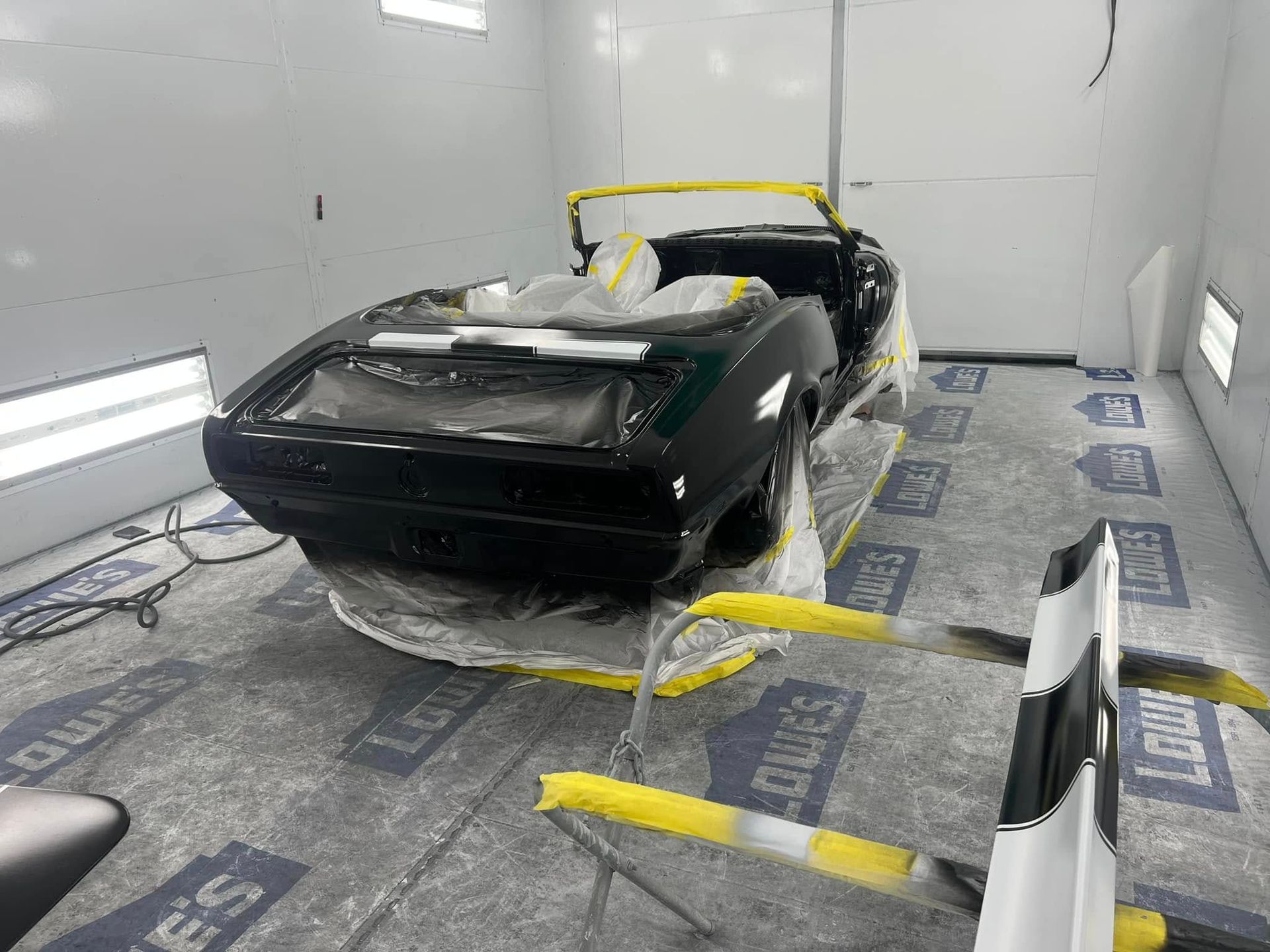 Black car partially masked with plastic and tape in a white paint booth, with rear end visible.