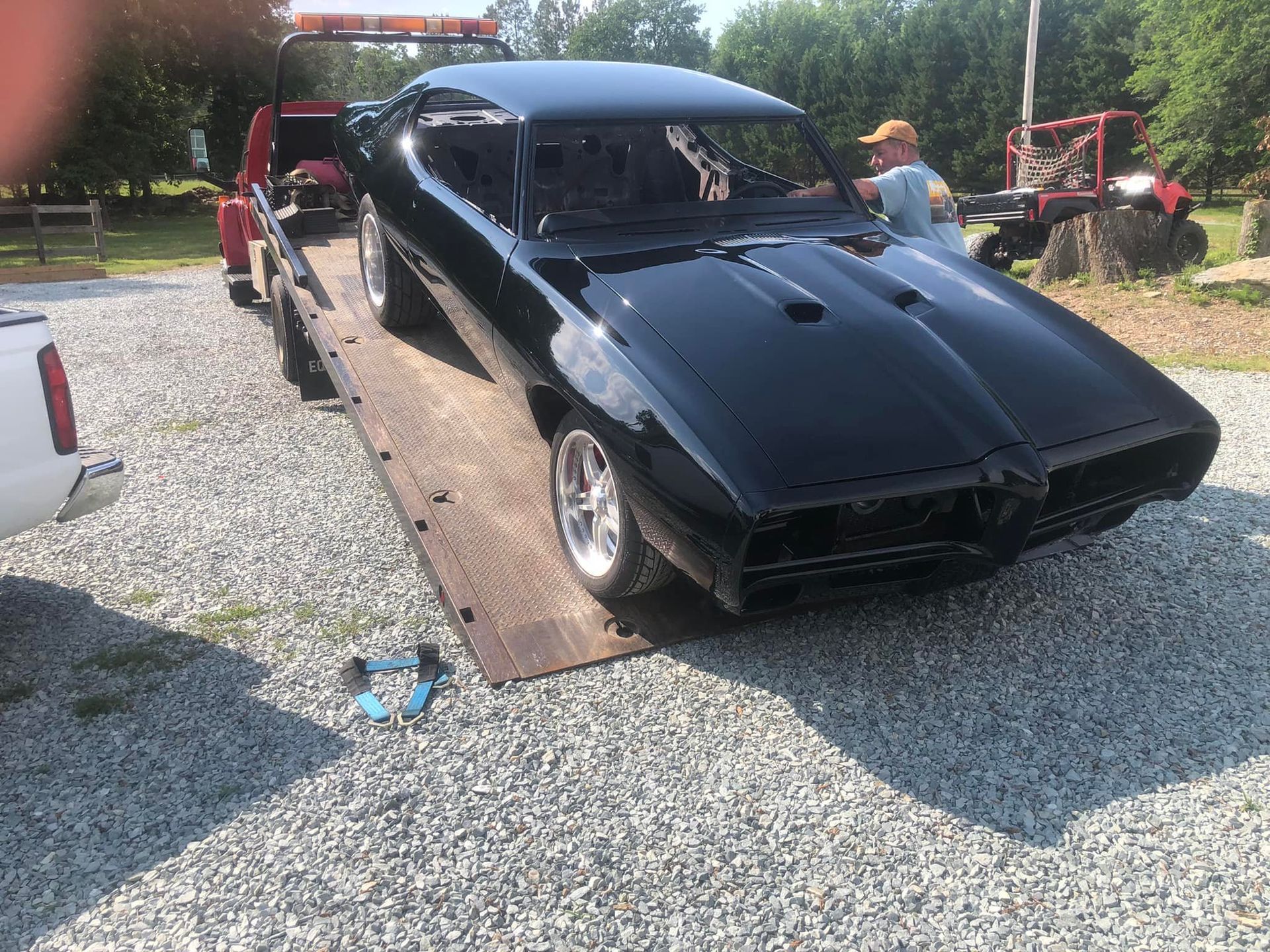 Black classic car on a trailer being towed in a gravel lot, with people working nearby.