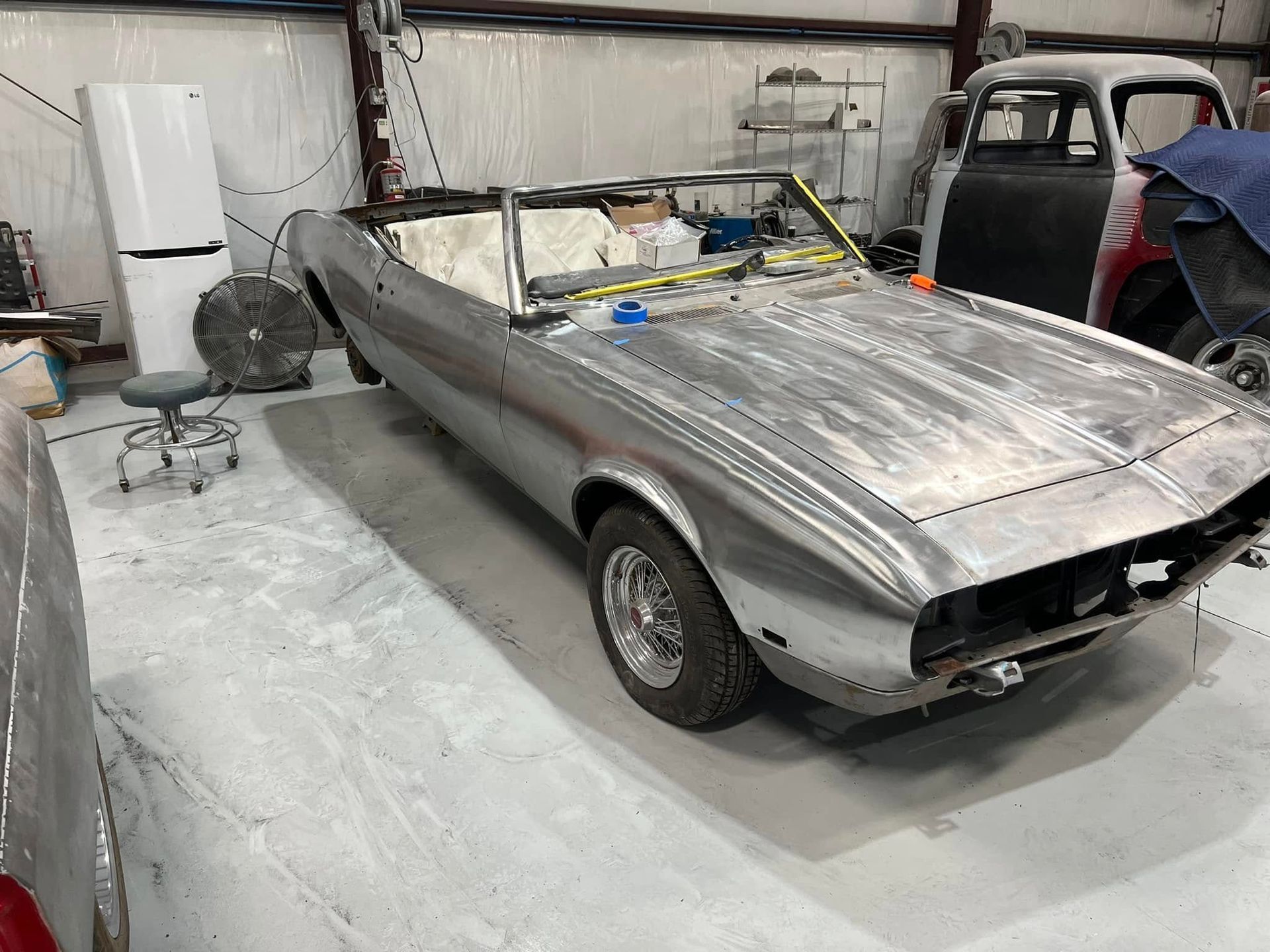 Silver classic convertible car under repair in a garage, with its hood and interior covered in protective plastic.