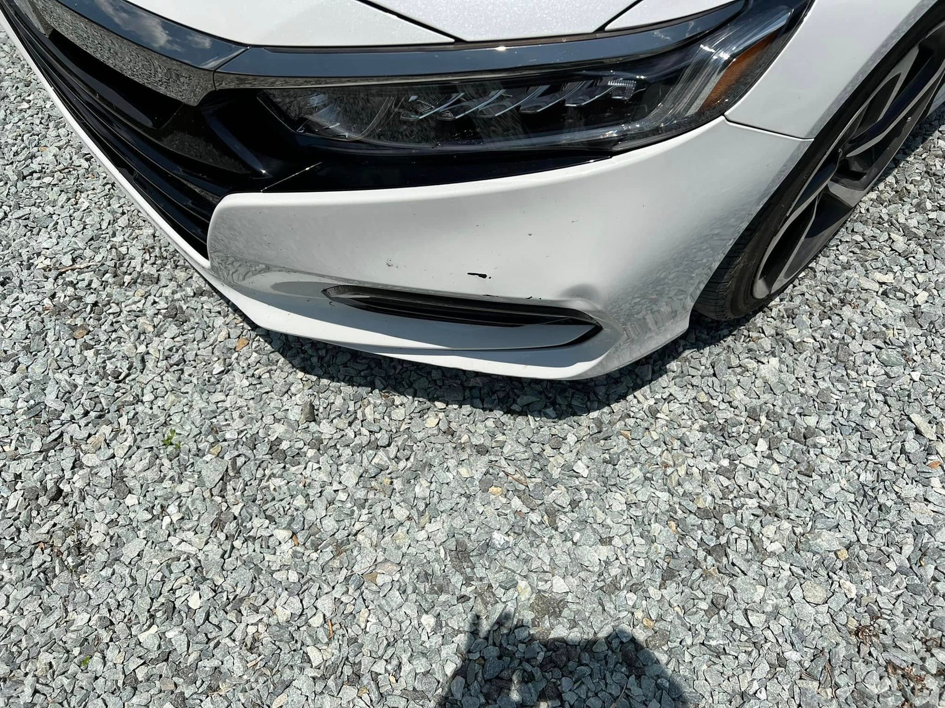 Front of a silver car with a damaged bumper, parked on gravel.