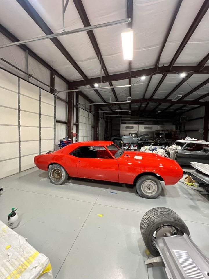 Red classic muscle car parked inside a large garage with other vehicles and equipment nearby.