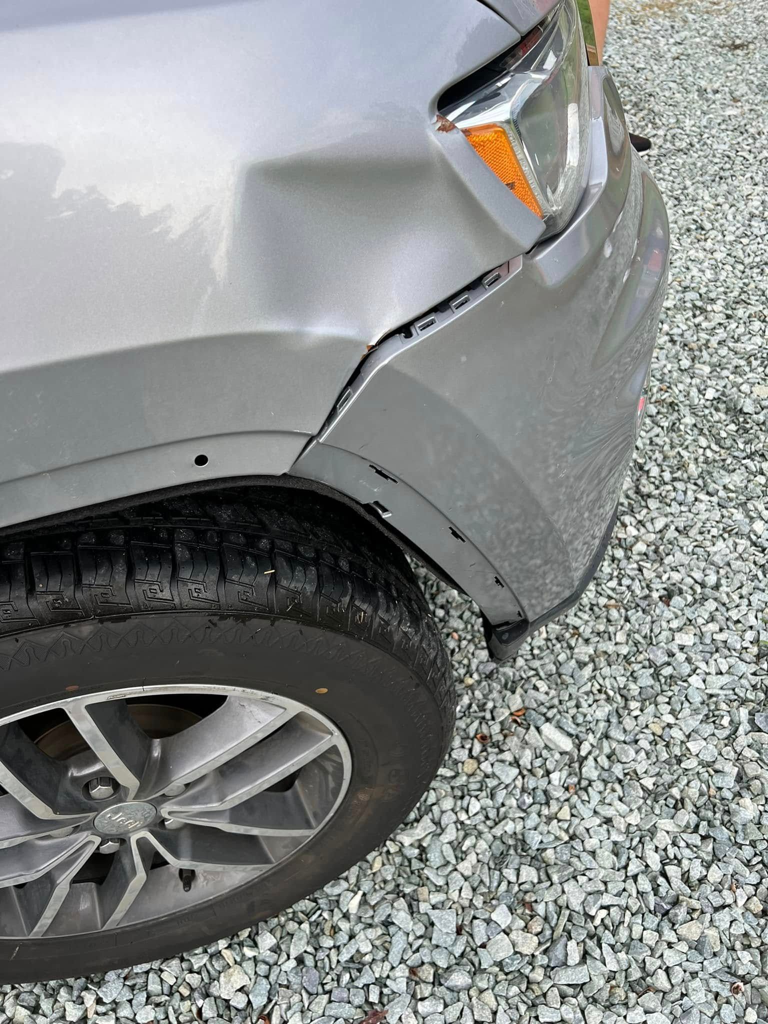 Close-up of a silver car’s front fender and wheel with a dented bumper on gravel.