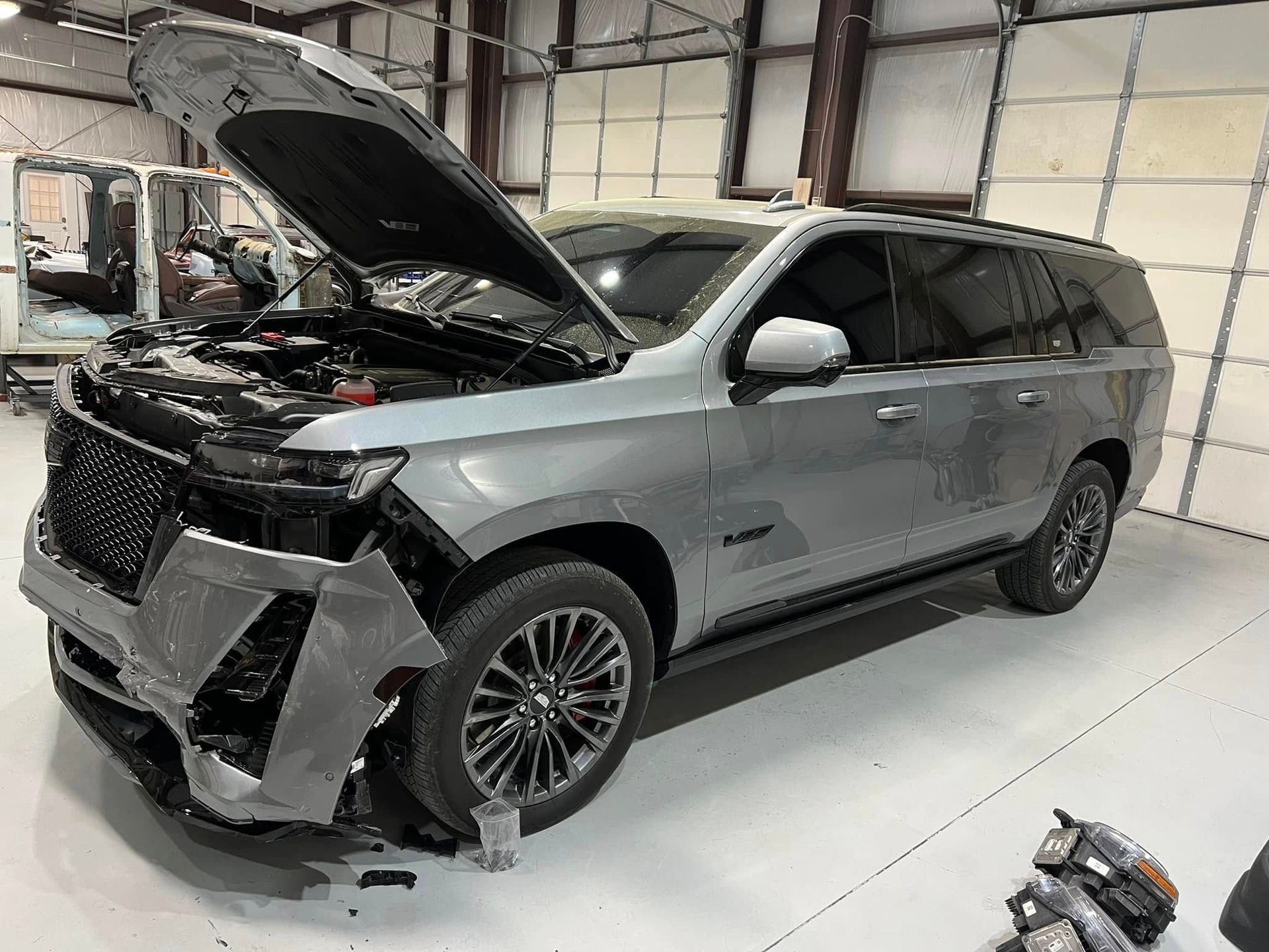 Silver SUV with front-end crash damage in a garage, hood open and bumper hanging off
