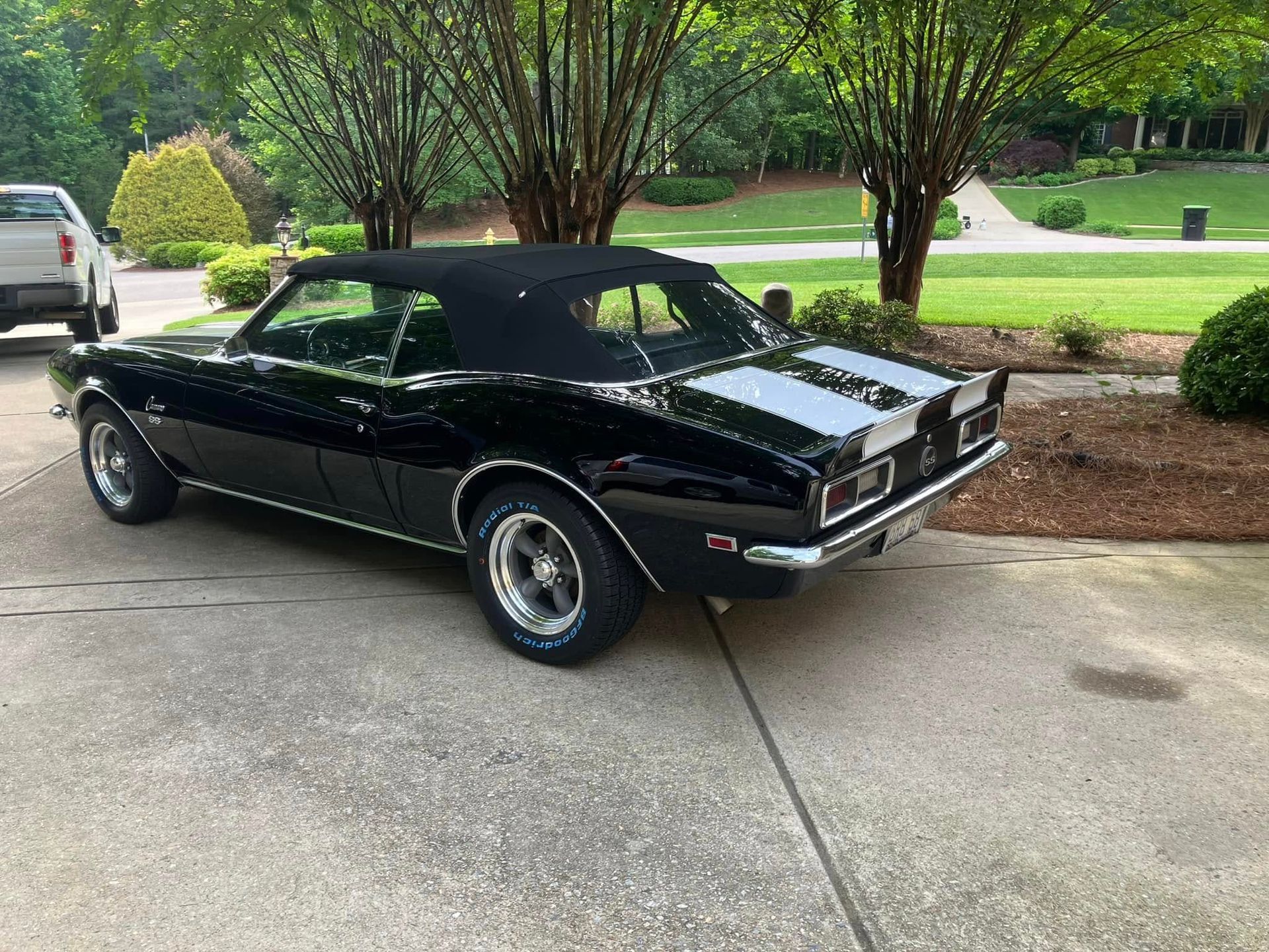 Black classic muscle car parked on a driveway under trees, side view with chrome wheels and white racing stripes