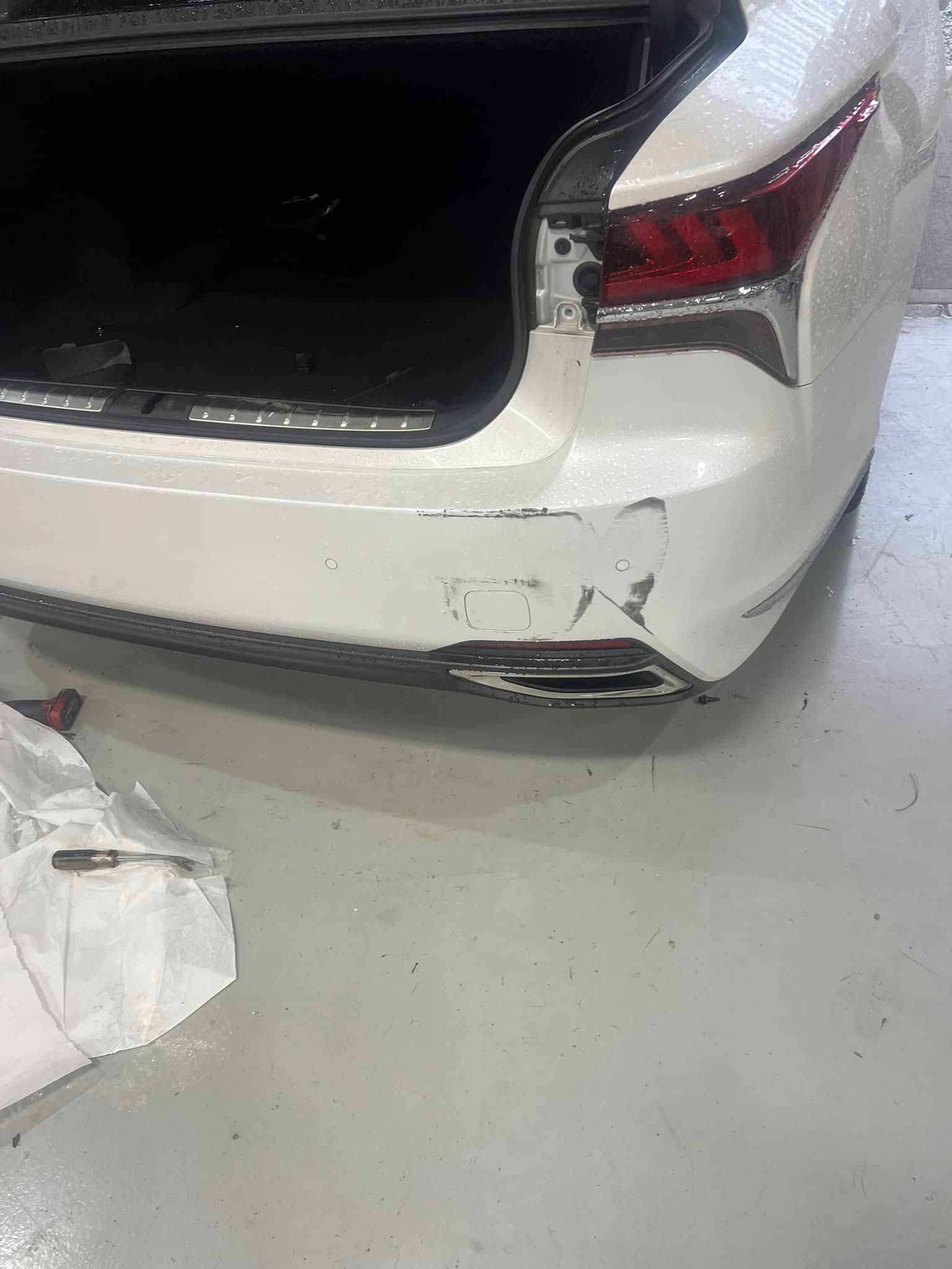 White car with rear bumper damage and scratches in a garage, trunk open.