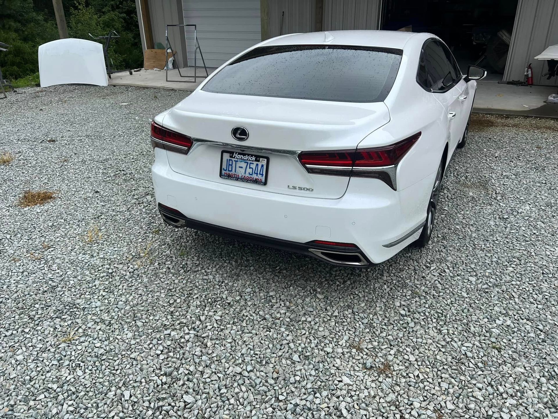 White sedan parked on gravel in a driveway, rear view with house and open garage in the background
