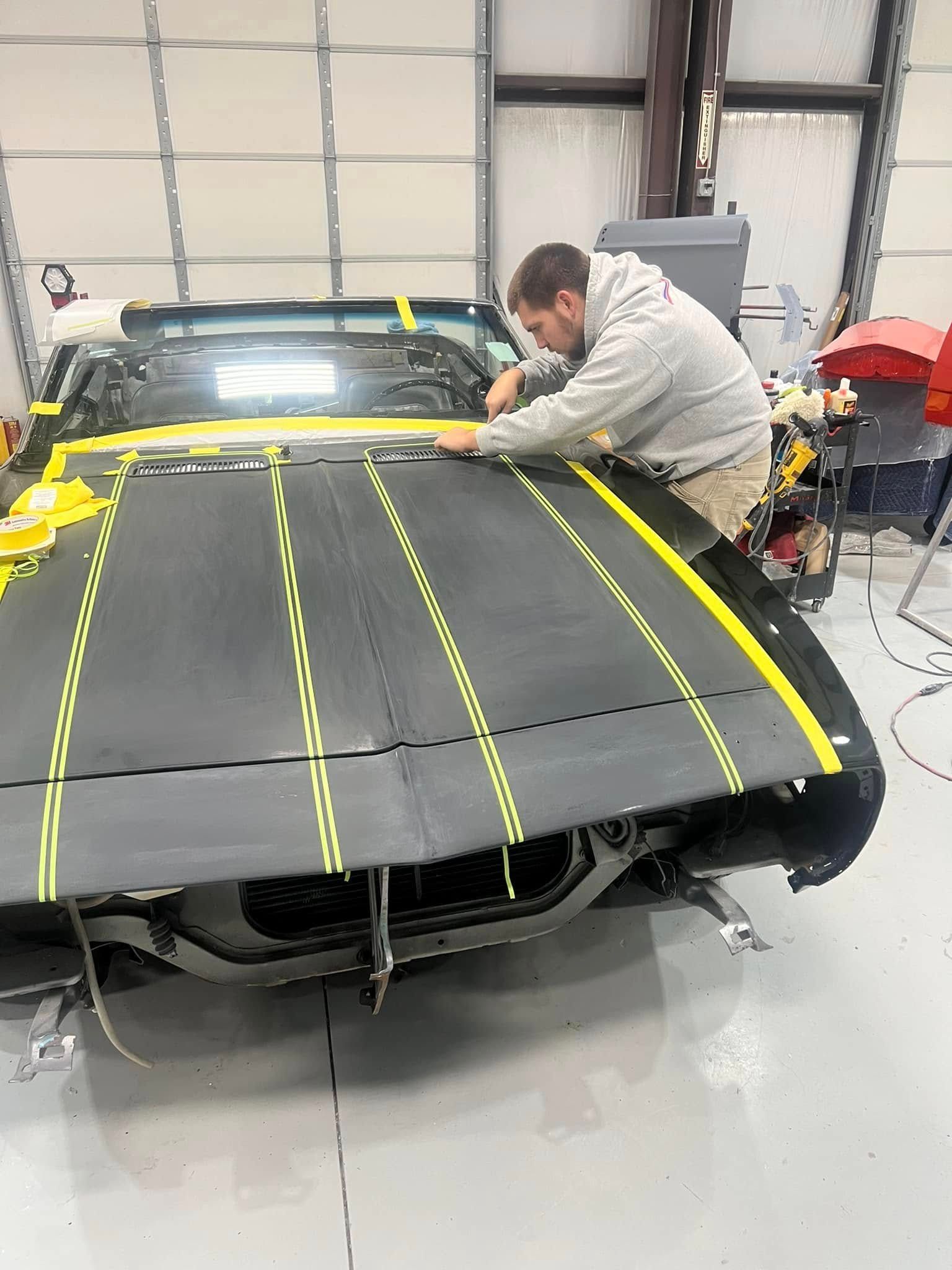 Man taping a matte black car hood with yellow masking tape in a garage workshop