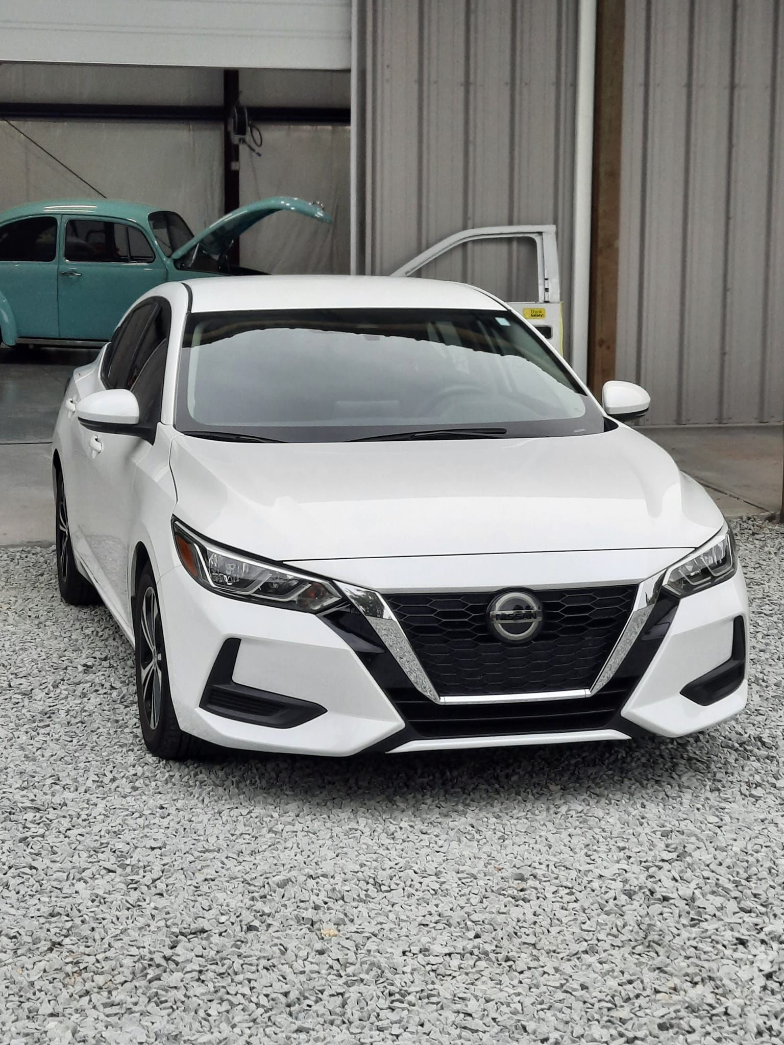 White Nissan sedan parked outside a building on a gravel lot, front view with a covered hood.