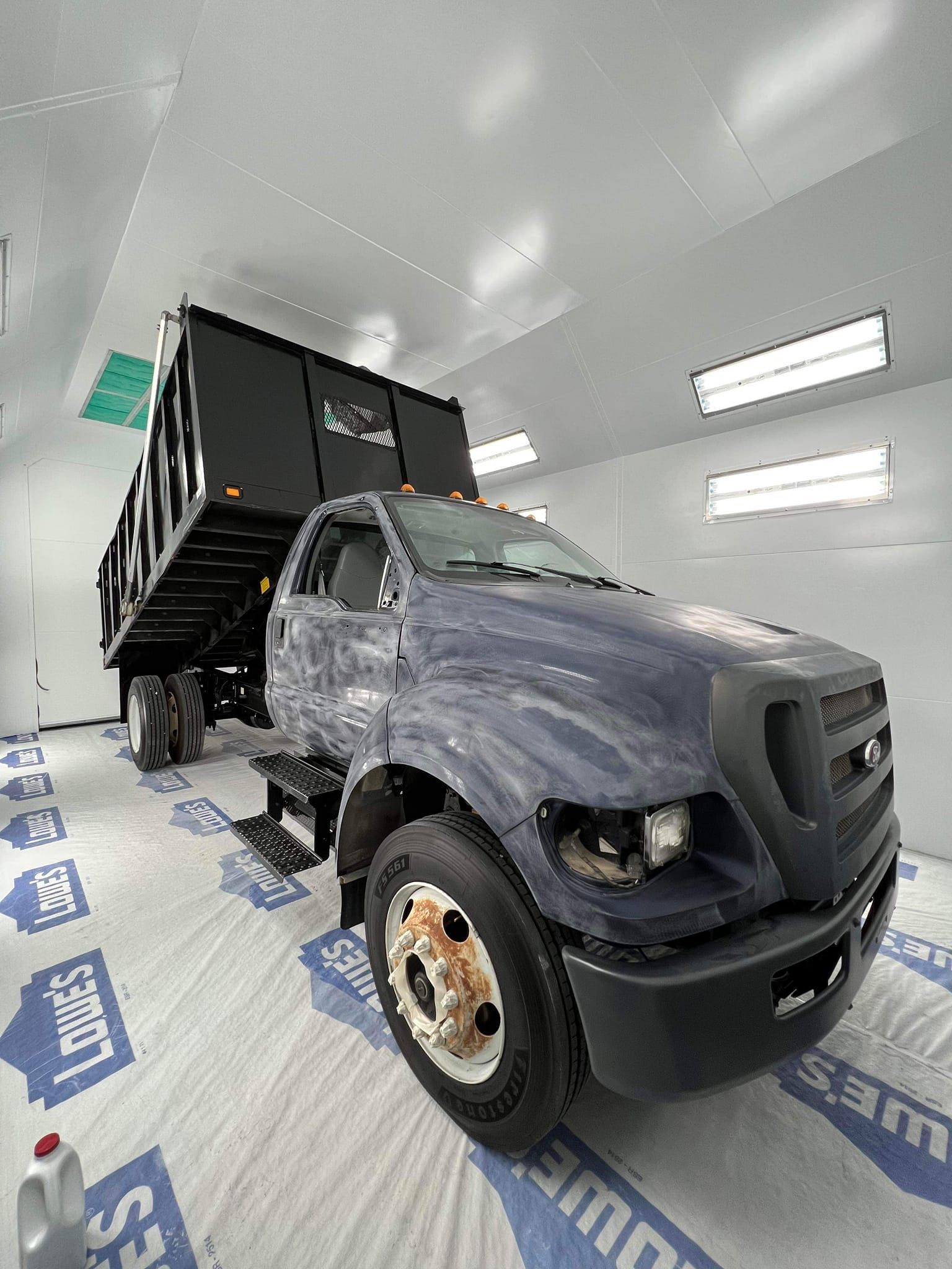 Gray dump truck inside a white paint booth with plastic-covered floor and ceiling lights