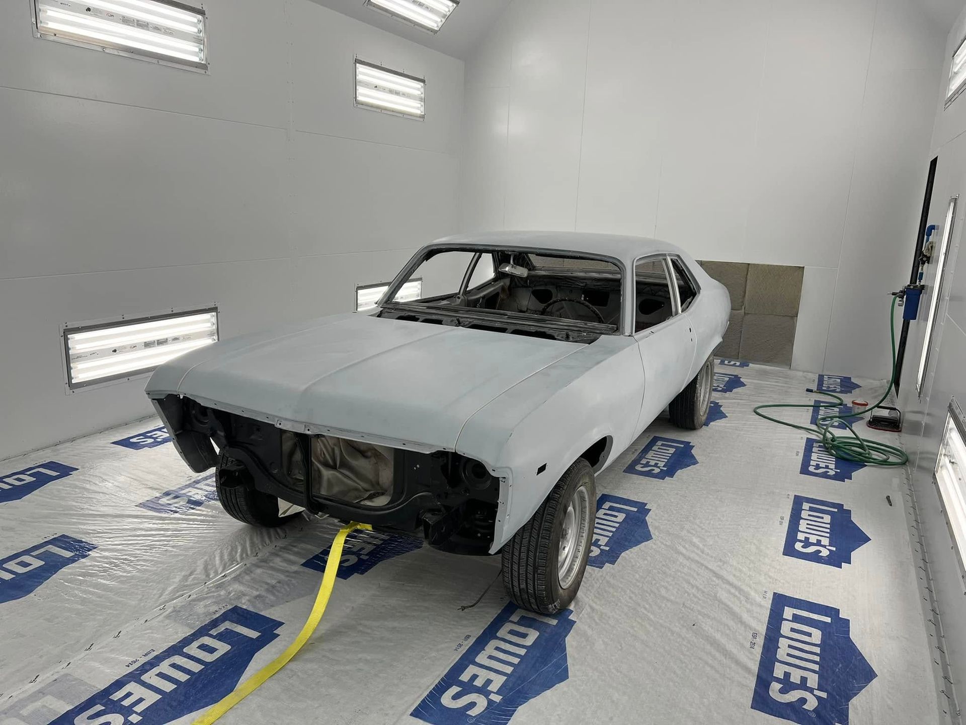 Primed white coupe body in a workshop paint booth, missing wheels and front bumper.