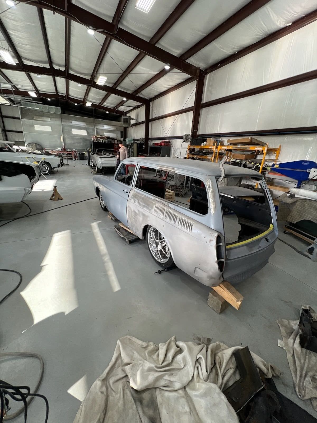 Partially restored silver station wagon in a large auto workshop with tools and cars nearby