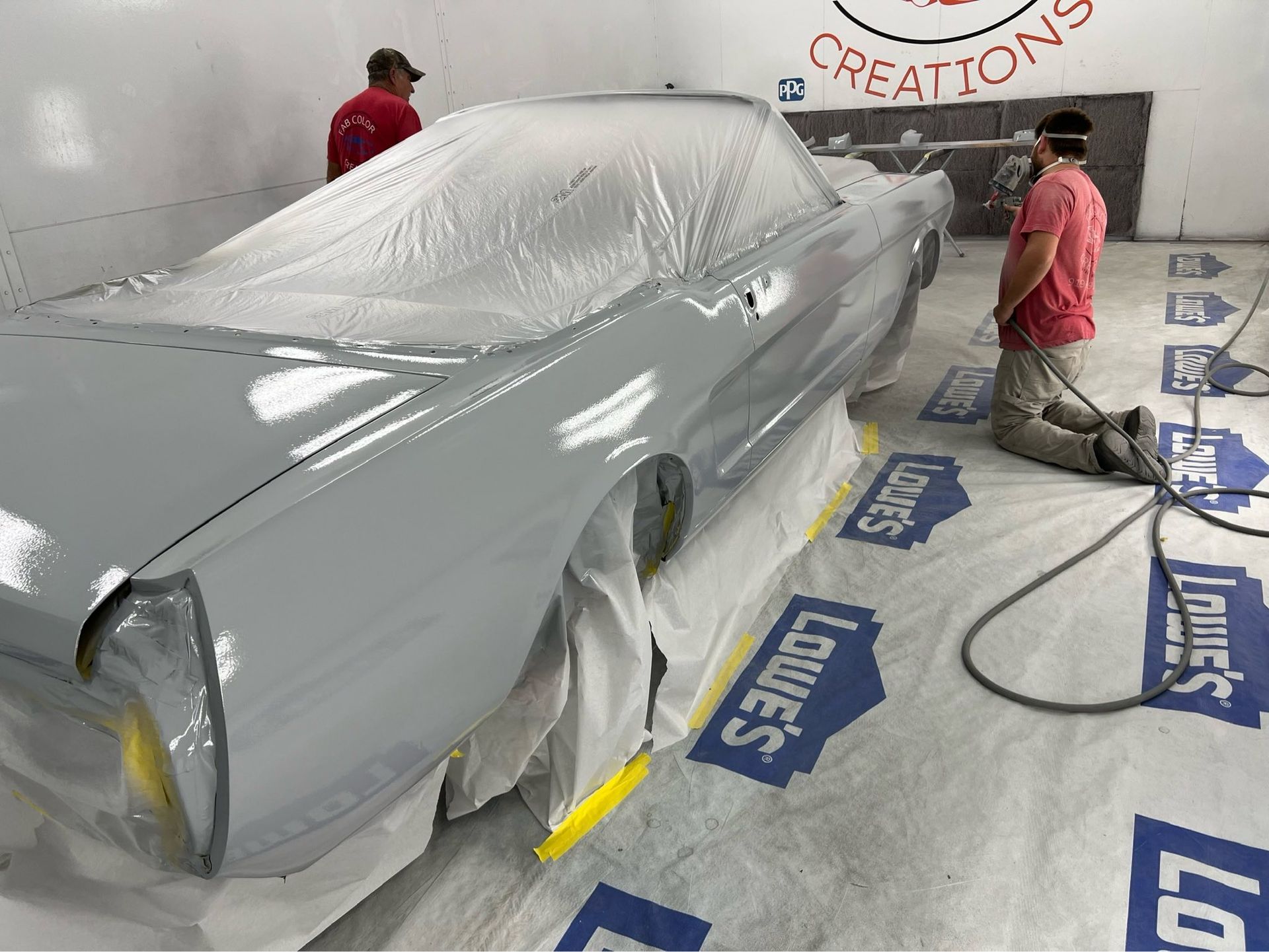 Two people spray-paint a silver car in a paint booth, with the vehicle masked in plastic.