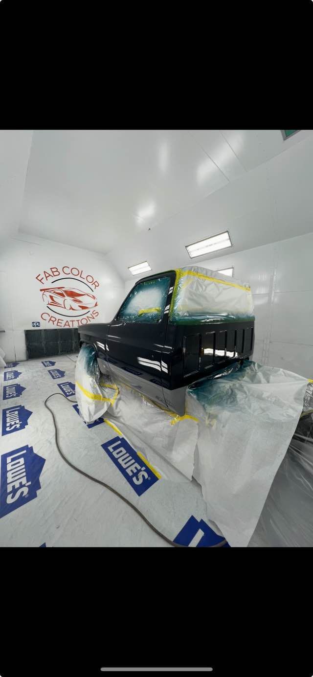 Black SUV in a bright white auto body spray booth, covered with protective plastic and tape