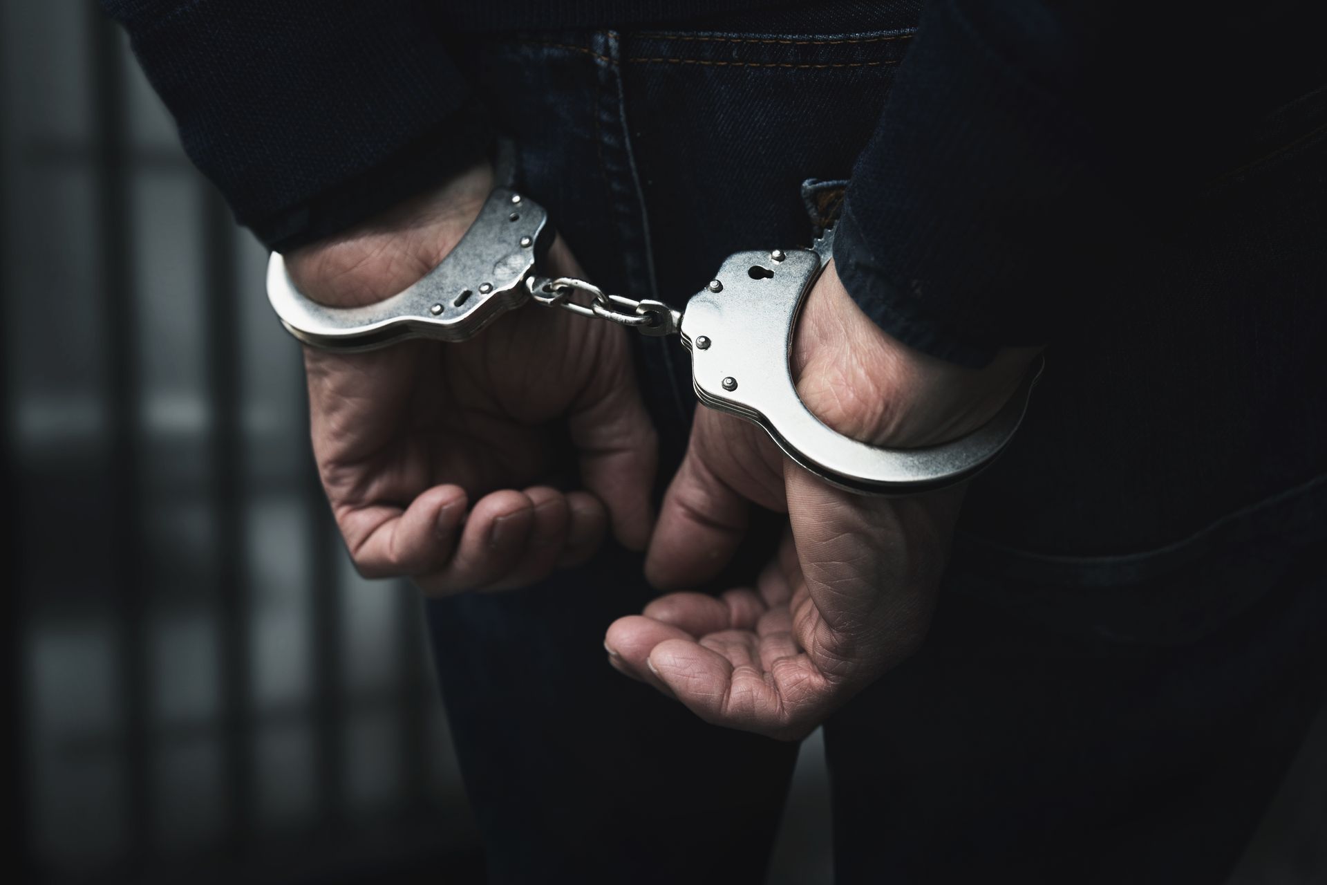 A person in dark clothing wearing metal handcuffs behind their back, with prison bars visible in the background.