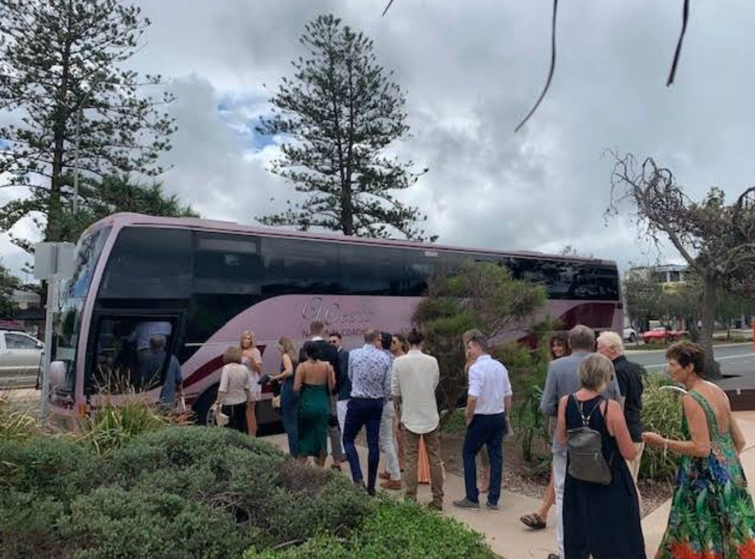 People gathered around a pink double-decker bus parked near greenery, under a cloudy sky.