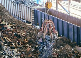 Grúa de garra industrial clasificando chatarra en un montón de escombros debajo de una cinta transportadora.