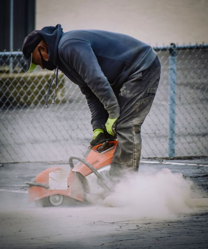 Worker using a concrete saw, cutting pavement and kicking up dust outdoors.