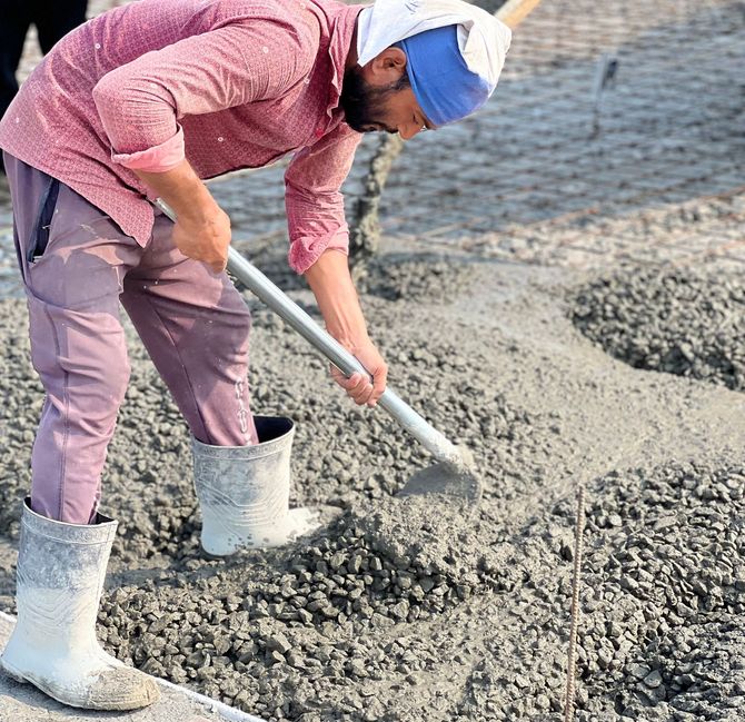 Worker shoveling wet concrete at a construction site, wearing boots and a cap.