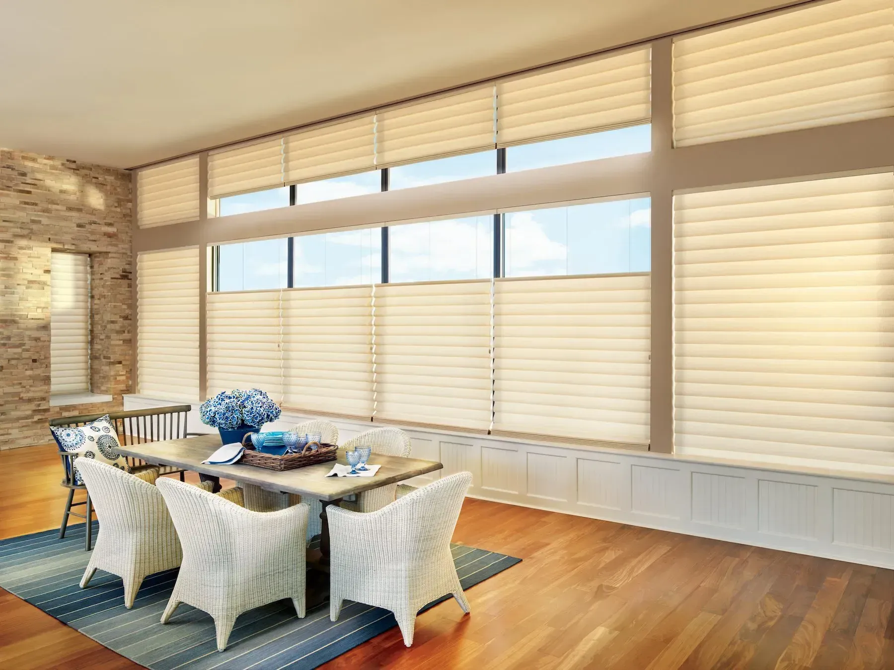 Bright dining room with wicker chairs, a table setting, wood floors, and large windows with blinds