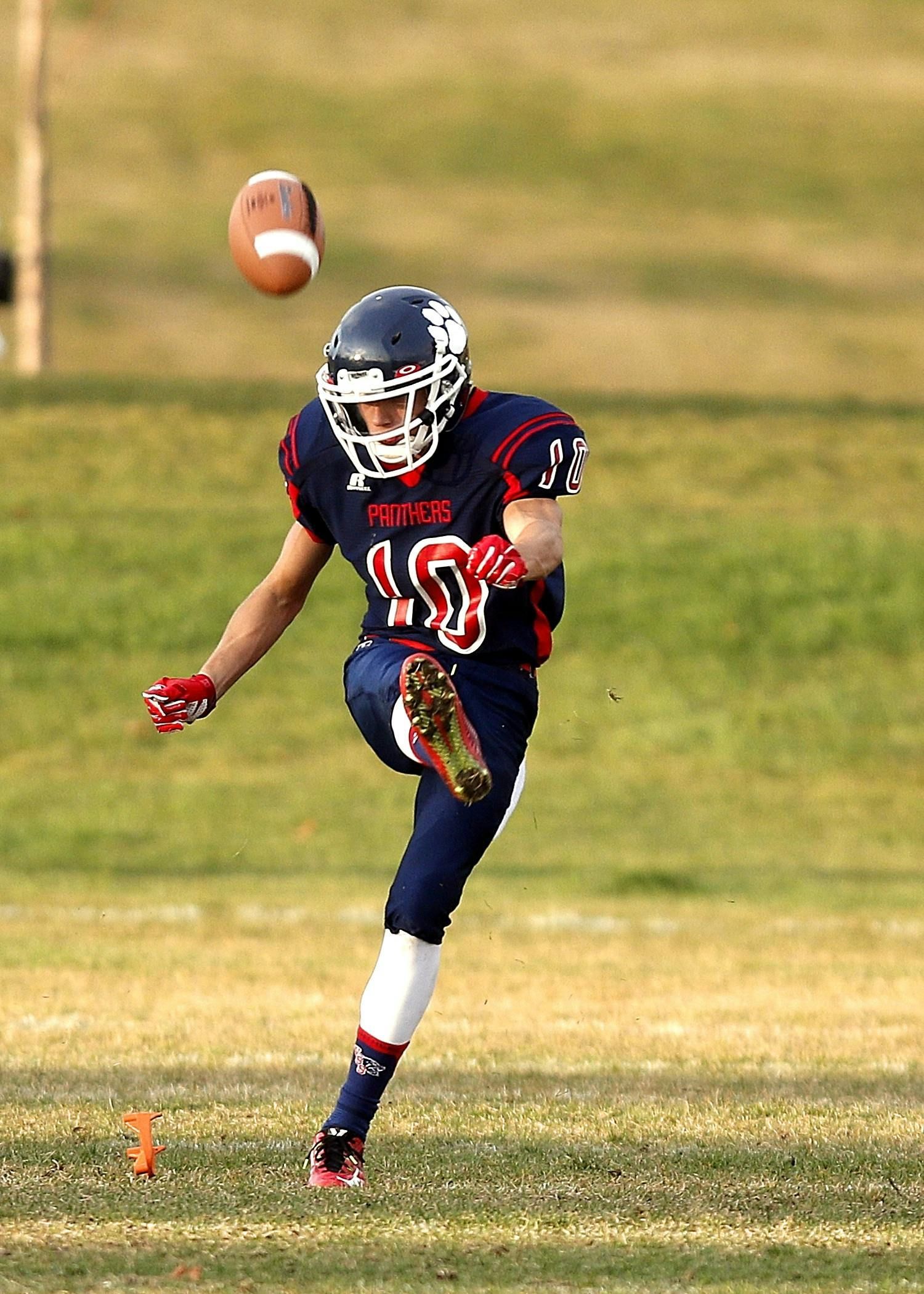 A football player in a dark blue uniform punts a football high into the air on a grassy field.