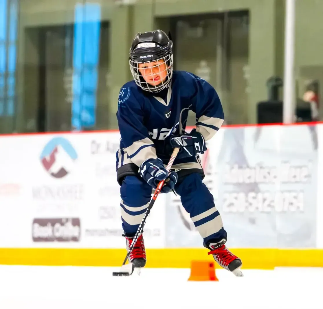 A young ice hockey player in a navy and grey uniform practices puck handling on an indoor rink.