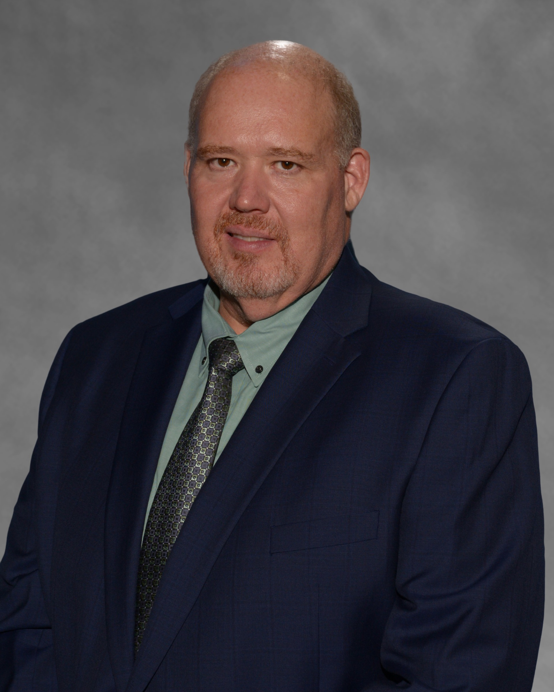 Man in a navy suit and green shirt, posing against a gray studio backdrop.