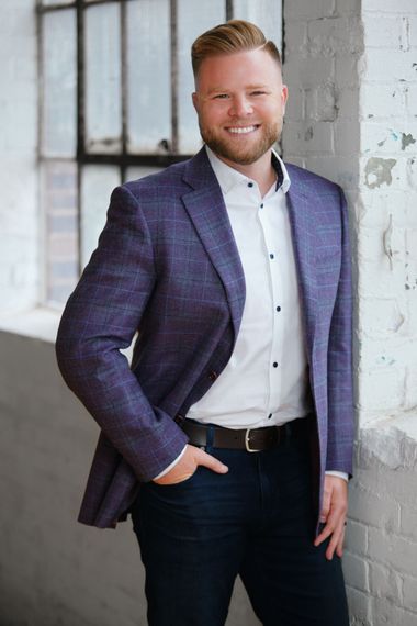 Smiling man in a navy blazer leaning against a white brick wall by a window.