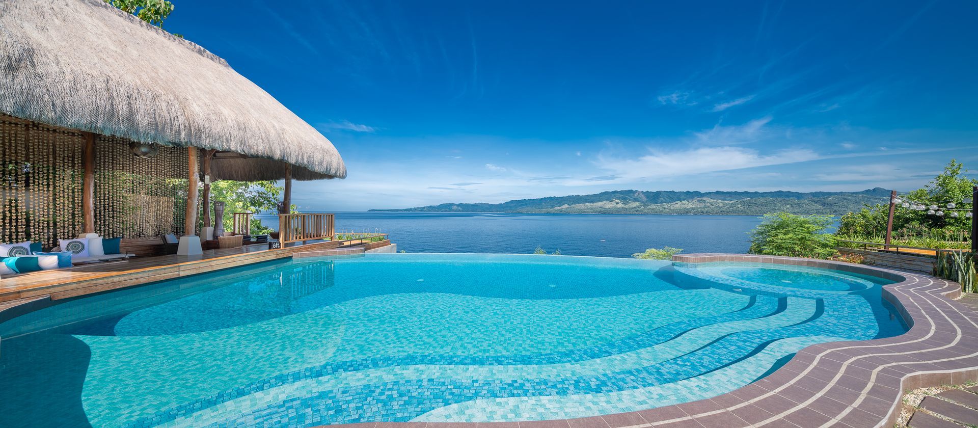 An infinity pool overlooks a body of water under a clear blue sky. A thatched roof shelter sits by the pool.