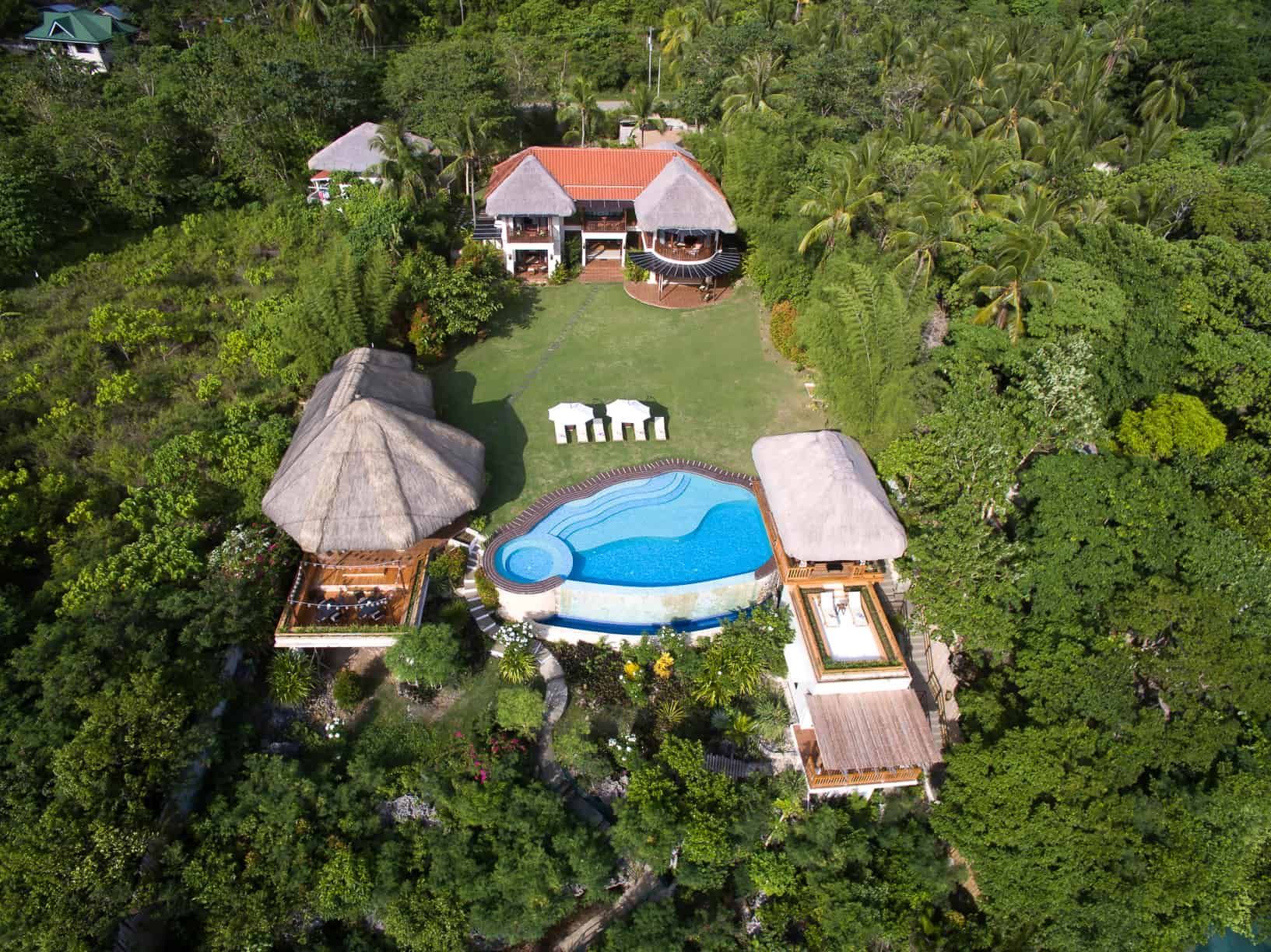 Aerial view of a tropical villa with a pool, grassy lawn, and thatched roof structures surrounded by lush greenery.