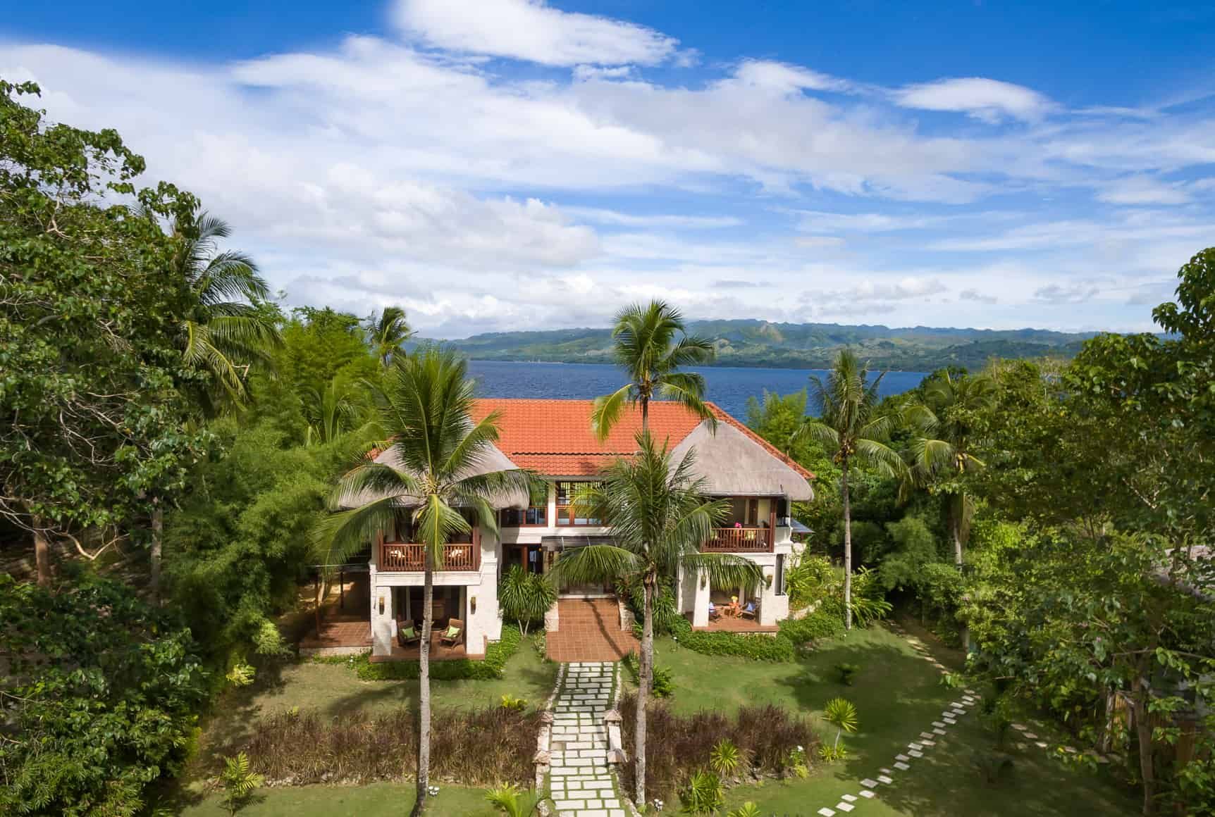Lush tropical landscape with a two-story house, red tile roof, and waterfront view under a blue sky.