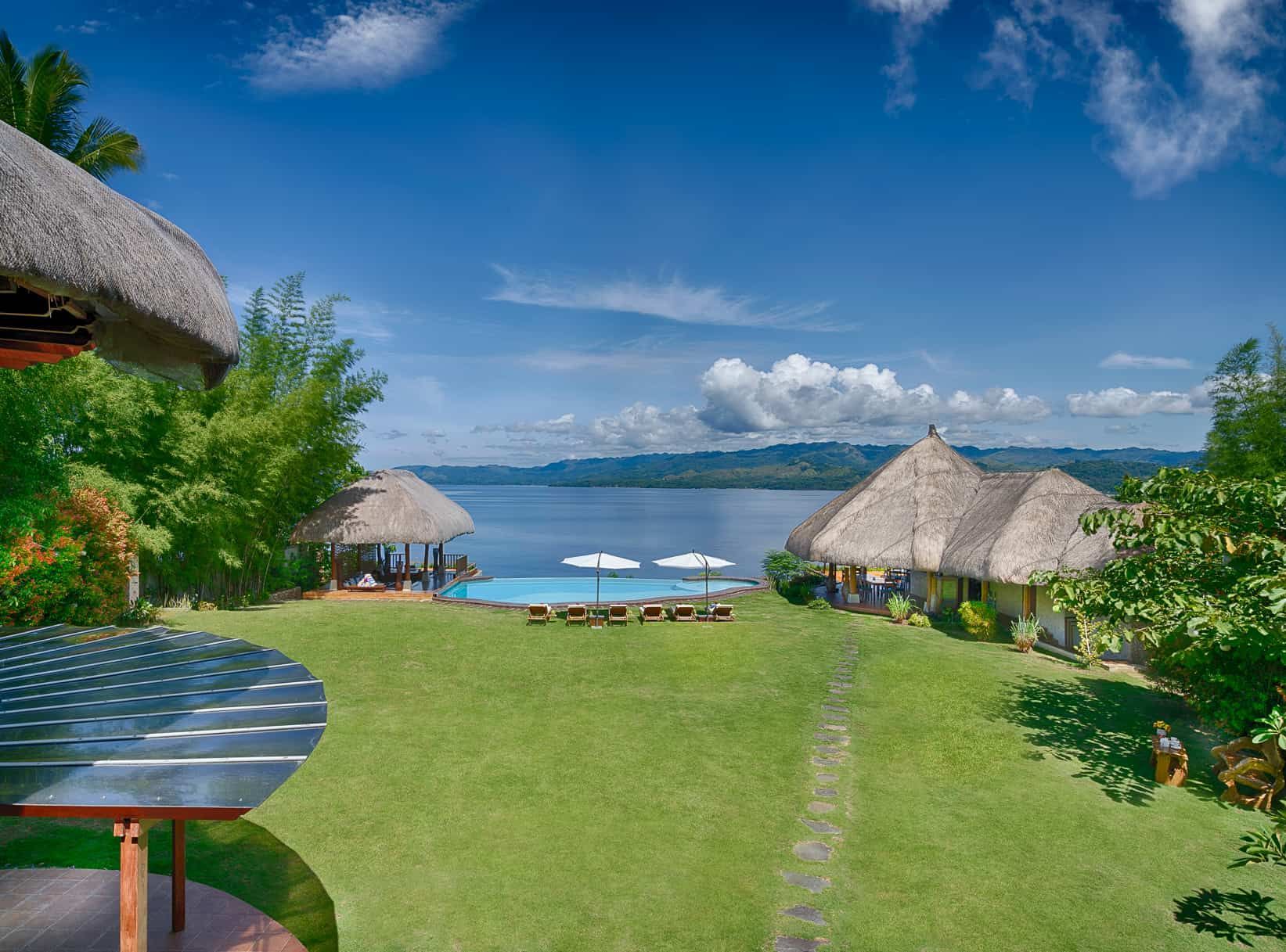 Lush green lawn with thatched-roof structures, a pool, and ocean view on a sunny day.
