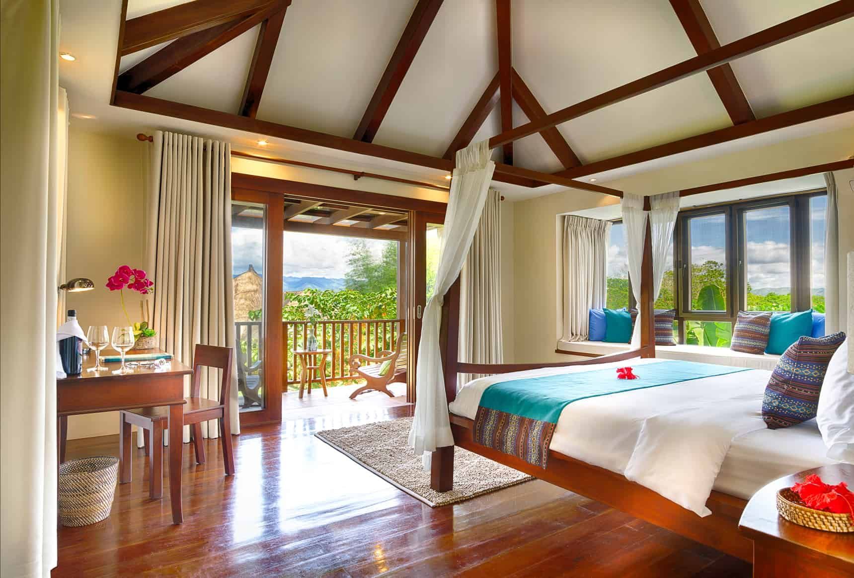 Bedroom with balcony: wooden four-poster bed, ocean view, desk with flowers, cream curtains, and brown hardwood floor.