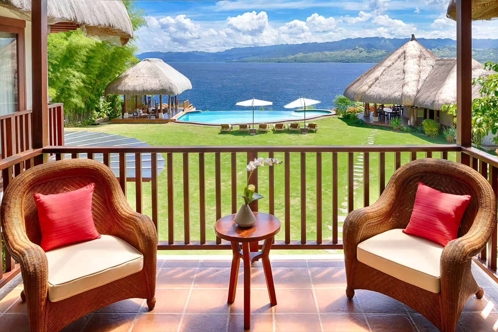 Balcony with wicker chairs, overlooking pool, ocean, and thatched-roof structures on a sunny day.