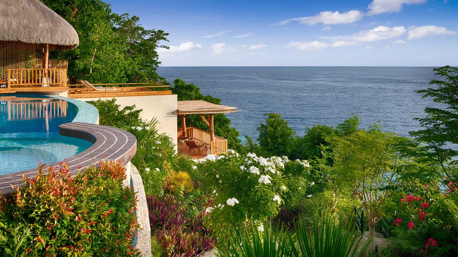 Infinity pool overlooks the ocean, lush greenery, and a thatched-roof structure.