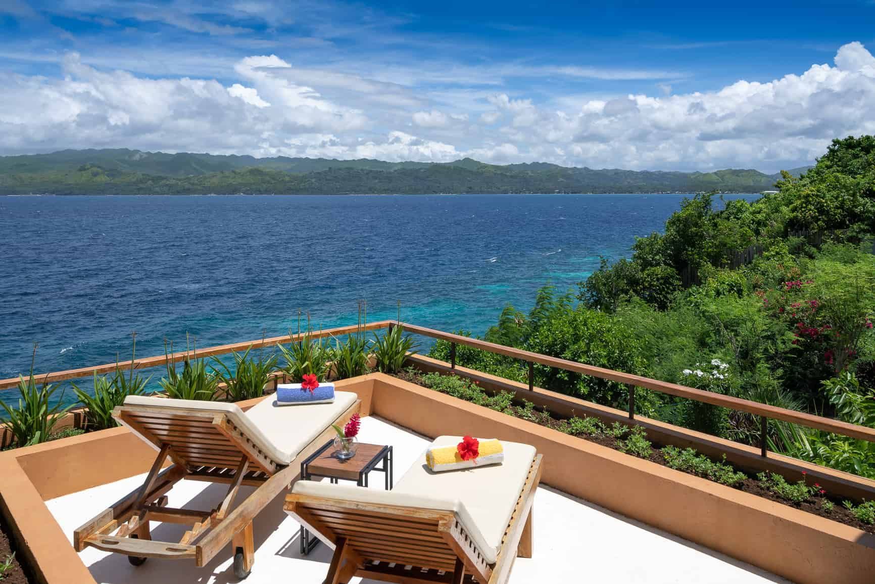 Two lounge chairs on a deck overlooking a blue ocean. Lush green vegetation and cloudy sky in the background.