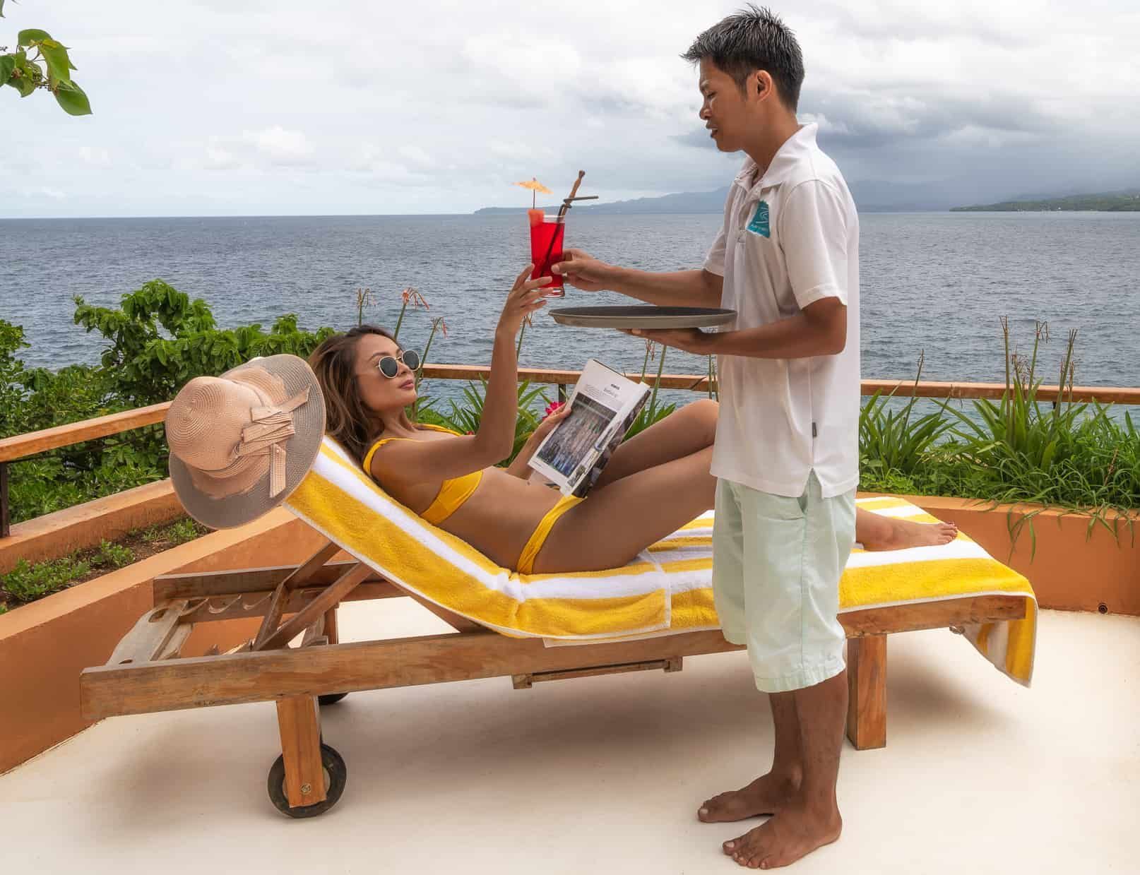 Woman on lounge chair receives cocktail from waiter, ocean view.