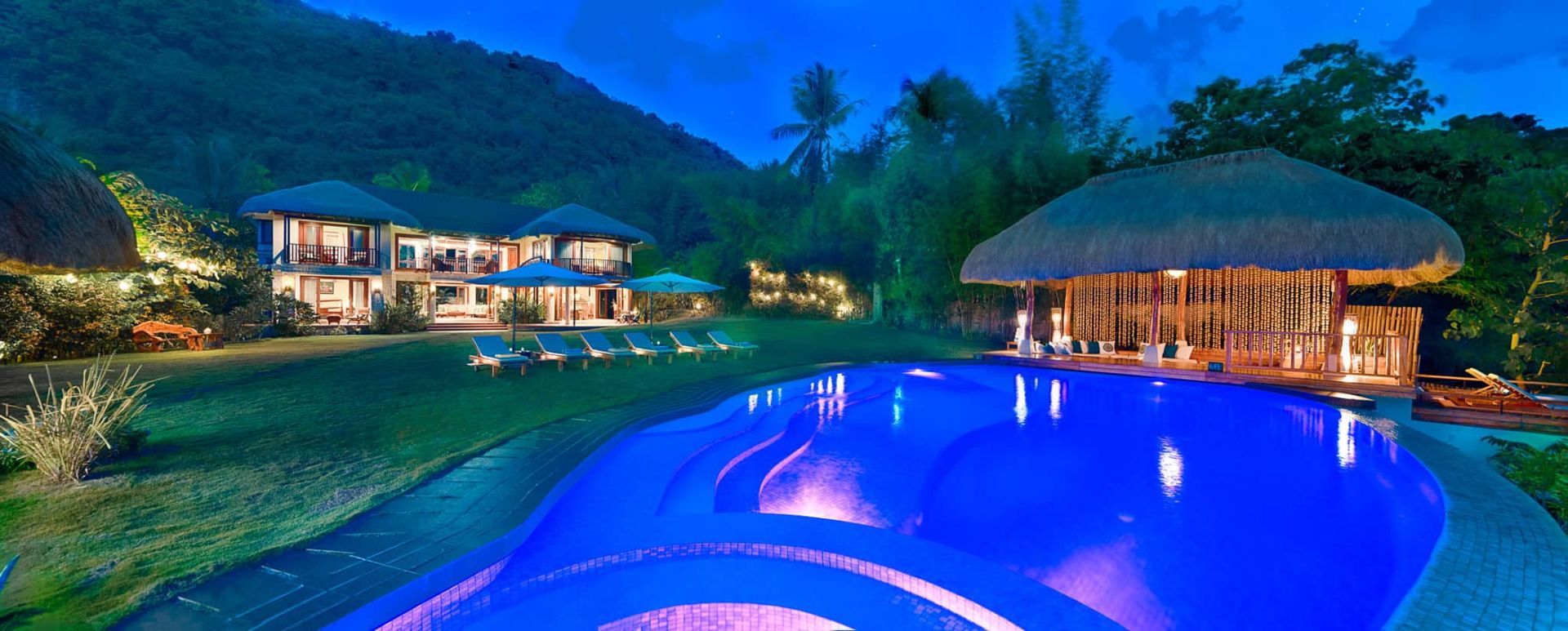 Night view of a resort with a pool, bar, and buildings surrounded by lush greenery and mountains.